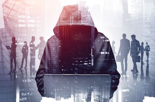 Silhouette of Hacker in a hoodie, sitting down and typing on laptop, against a background showing silhouettes of business professionals standing